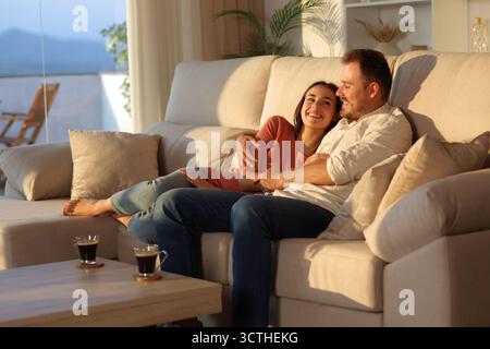 Glückliches Paar, das sich bei Sonnenuntergang zuhause auf einer Couch sieht Stockfoto