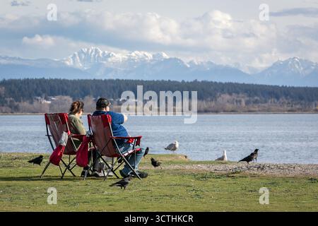 Mann und Frau sitzen draußen in Campingstühlen am sonnigen Herbsttag, Blick nach hinten. Ein Paar sitzt auf den Klappstühlen in der Nähe des Sees oder des Meeres, genießt die Ruhe Stockfoto