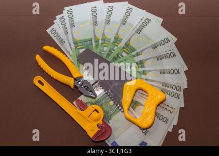 Satz gelber Spielzeugwerkzeuge, einschließlich Säge, Schraubenschlüssel und Zange, die auf einem Fächer von 100-Euro-Banknoten platziert werden. Das Konzept der finanziellen Reparatur, der wirtschaftlichen Kontrolle und der Mononie Stockfoto