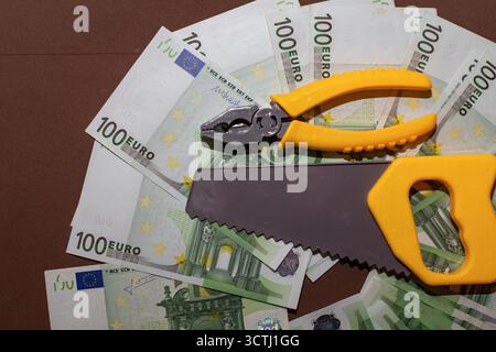 Satz gelber Spielzeugwerkzeuge, einschließlich Säge, Schraubenschlüssel und Zange, die auf einem Fächer von 100-Euro-Banknoten platziert werden. Das Konzept der finanziellen Reparatur, der wirtschaftlichen Kontrolle und der Mononie Stockfoto