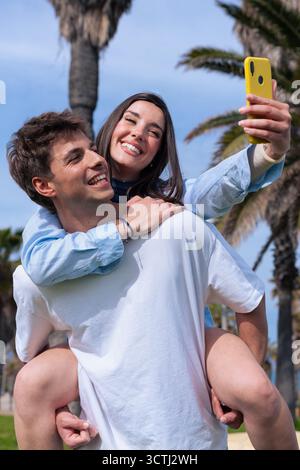Glückliches Paar, das Selfie mit dem Smartphone macht, während es am Strand mit dem Huckepack telefoniert Stockfoto