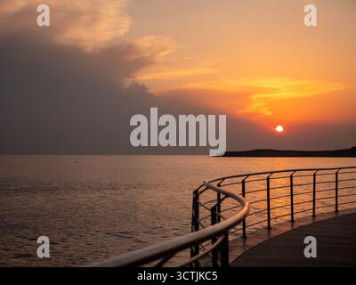 Orange Kugel eines Sonnenuntergangs über dem Mittelmeer in Ayia Napa, Blick von der modernen Promenade mit Metallgeländer über kurviges Ufer und Sonnenuntergang Stockfoto