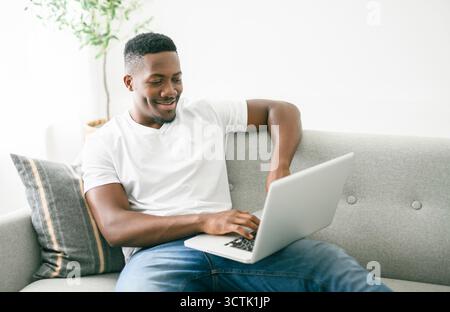 Fröhlicher schwarzer Mann, der den Computer auf dem Sofa benutzt. Stockfoto