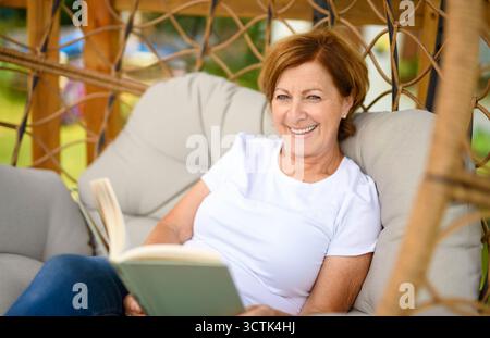 Ein Porträt einer Frau mittleren Alters, die Buch auf Hamacstuhl liest Stockfoto