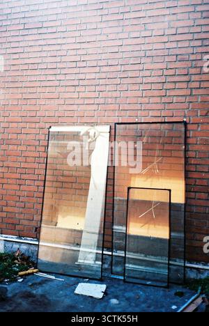 Fensterscheiben, die an einer roten Ziegelwand gestapelt sind, wurden analog in einem vertikalen Rahmen aufgenommen, mit hellen Sonnenreflexionen auf dem Glas. Stockfoto