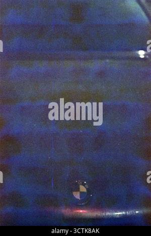 Analoge genarbte Nachtaufnahme einer schwarzen BMW-Kapuze mit verschwommenen Blautönen in den dunklen Bereichen, die eine abstrakte, stimmungsvolle Komposition schaffen. Stockfoto