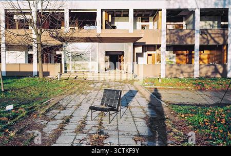 Leerer Stuhl in der frühen Wintersonne vor einer verlassenen Fabrik, mit dem Schatten des Fotografen im Hintergrund, aufgenommen auf analogem 35-mm-Film. Stockfoto