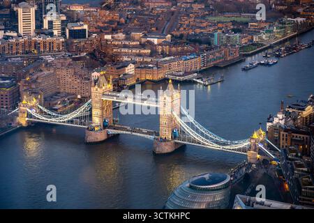 Ein atemberaubender Blick aus der Vogelperspektive fängt die Tower Bridge in der Abenddämmerung ein, unter der die Themse fließt und die Lichter der Stadt im Wasser reflektieren, was Londons lebhafte Abendatmosphäre unterstreicht. Stockfoto