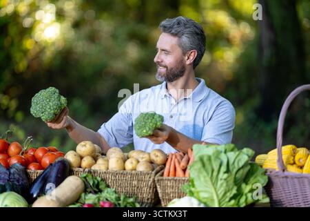 Landwirt, der frisches Gemüse im ländlichen Garten erntet. Mann überprüft Kürbisse, Karotten, Mais, Brokkoli, Paprika, Tomaten und Kartoffeln im Freien Stockfoto