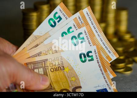 Nahaufnahme einer Hand, die mehrere 50-Euro-Banknoten mit Goldmünzstapeln im Hintergrund hält, die Finanzen, Vermögen, Ersparnisse und Investitionen symbolisieren Stockfoto