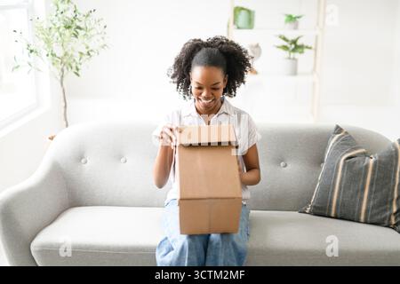 Glückliche schwarze Dame erhielt das Paket, packte den Karton aus und saß auf dem Sofa. Stockfoto