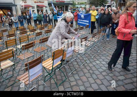 Flensburg, Schleswig-Holstein Flensburg gedenkt den Opfern des 7. Oktober. Eine Frau befestigt Papierbögen an Stühlen auf einem Marktplatz, im Hintergrund stehen Menschen mit Transparenten und Israelfahnen. Es scheint eine Gedenk- oder Solidaritätsveranstaltung zu sein, zahlreiche leere Stuhlreihen sind auf dem Kopfsteinpflaster aufgebaut. Die Versammlung wirkt ruhig und organisiert. Aufnahme vom 07.10.2025, Flensburg, Südermarkt. *** Flensburg, Schleswig Holstein Flensburg gedenkt an die Opfer des 7. Oktober Eine Frau klebt auf einem Marktplatz Papierbögen auf Stühlen, im Hintergrund AR Stockfoto