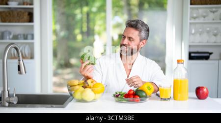 Ein reifer Mann frühstückt in der Küche mit frischem Müsli und Milch. Man isst gesundes Mittagessen genießt einen nahrhaften Salat und Gemüse zu Hause. Leckerer Snack Stockfoto