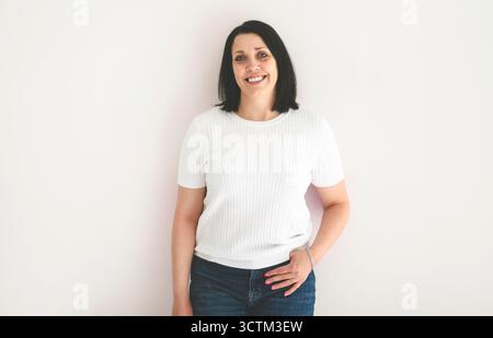 Echte und authentische reife Frau, die in die Kamera blickt und lächelt, während sie vor weißem Hintergrund steht Stockfoto