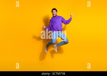 Der energiegeladene junge Mann mit violettem Hoodie springt fröhlich auf leuchtend gelbem Hintergrund und drückt Aufregung und Positivität aus Stockfoto