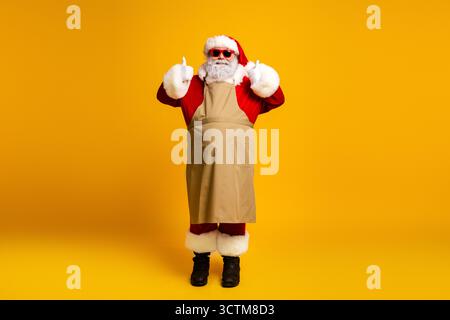 Der Weihnachtsmann, der als cooler Werkstattkoch gekleidet ist, posiert in festlicher Kleidung mit fröhlichem Ausdruck und Sonnenbrille Stockfoto