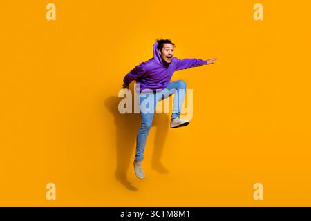 Energiegeladener junger Mann in einem violetten Hoodie, der vor einem leuchtend gelben Hintergrund springt und Freude und Begeisterung zum Ausdruck bringt Stockfoto