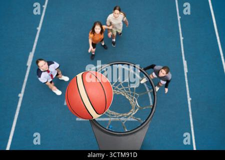 Eine Gruppe von vier Teenagern, darunter zwei kaukasische Mädchen und zwei Jungen verschiedener Ethnien, spielen im Freien Streetball, stehen auf dem blauen Platz und beobachten Basketball, der sich dem Basketball nähert Stockfoto