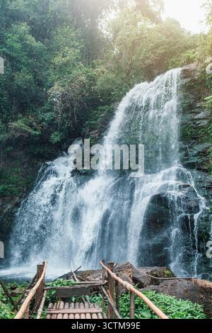 Mächtiger Wasserfall, der über zerklüftete Felsen in einen üppigen Pool mit Bambussteg stürzt, umgeben vom grünen Dschungel des Doi Inthanon National Park, Chiang Stockfoto