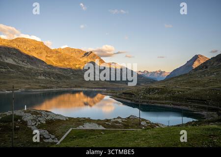 Der Lago Bianco schimmert unter der aufgehenden Sonne am Berninapass in Graubünden. Die umliegenden Berge reflektieren das ruhige Wasser und zeigen die natürliche Schönheit der Schweizer Alpen. Stockfoto