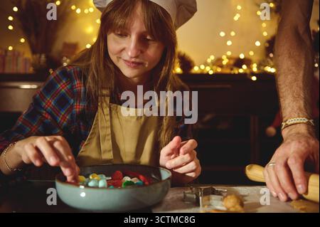 Ein Mädchen mit beigefarbener Schürze und Kochmütze ziert eine Schüssel mit bunten Süßigkeiten in einer warmen, festlichen Küche mit funkelnden Lichtern. Stockfoto