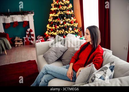 Frau in rotem Pullover, die sich während der festlichen Weihnachtsfeier in einem gemütlich dekorierten Wohnzimmer auf der Couch entspannt Stockfoto