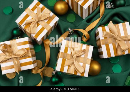 Fünf weiß-goldgestreifte Geschenkboxen mit goldenen Bändern auf grünem Seidenhintergrund. Luxuriöse Geschenkboxen, Weihnachtskugeln und rundes farbenfrohes Konfekt Stockfoto