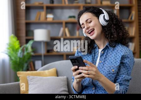 Junge Frau mit lockigem Haar, die auf einer Couch liegt, weiße kabellose Kopfhörer trägt und ein Smartphone hält, und fröhlich zur Musik mitsingt, während sie sich zu Hause entspannt Stockfoto
