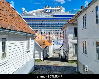 Historische weiß bemalte Holzhäuser auf einer schmalen Kopfsteinpflasterstraße mit einem Kreuzfahrtschiff im Hintergrund in Gamle Stavanger, Norwegen Stockfoto