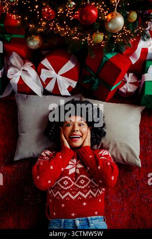 Fröhliche Frau in rotem Weihnachtspullover liegt unter einem festlichen Weihnachtsbaum, umgeben von bunten Geschenken im Weihnachtsambiente Stockfoto