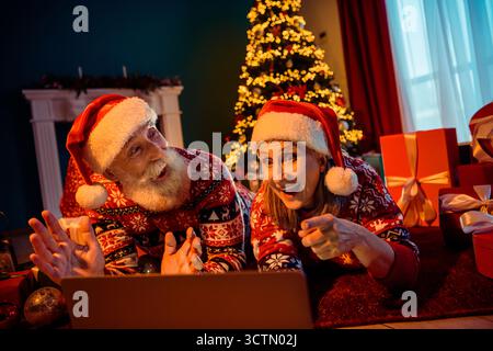 Glückliches Paar in Weihnachtsmützen Genießen Sie einen gemütlichen Weihnachtsabend zu Hause bei einem leuchtenden Baum mit Geschenken und festlichen Lichtern Stockfoto