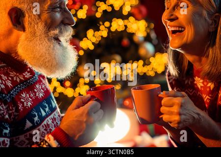 Warme Weihnachtsszene mit einem älteren Paar, das Kaffeetassen teilt und neben einem leuchtenden Weihnachtsbaum lacht Stockfoto