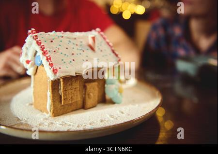 Ein dekoriertes Lebkuchenhaus sitzt auf einem schneebedeckten Teller, Zuckerstaub, farbenfrohe Süßigkeiten und weiche Weihnachtslichter im Hintergrund, die warme Familie CH einfangen Stockfoto