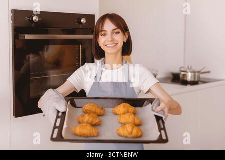 Glückliche junge Hausfrau hält Tablett mit frisch gebackenen Croissants, steht in der Küche und lächelt vor der Kamera Stockfoto