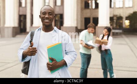 Glücklicher afroamerikanischer Student, der draußen mit Klassenkameraden auf dem Universitätscampus steht, posiert und vor der Kamera lächelt Stockfoto