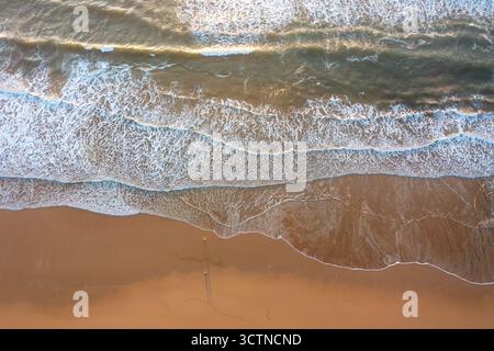 Drohnenaufnahme von Meereswellen, die auf den Sandstrand stürzen. Weiße Schaummuster bilden sich, wenn Wasser auf das Ufer trifft. Sand erscheint in der Nähe der Wasserlinie glatt Stockfoto