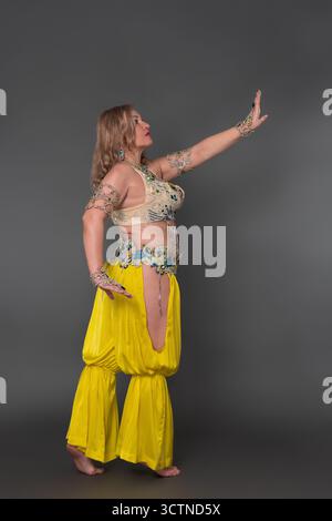 Kurvige Tänzerin mit handerhöhter Kleidung im Tanzoutfit - Oberteil und gelbe Haremshosen und ohne Schuhe oder Socken, in voller Länge Stockfoto
