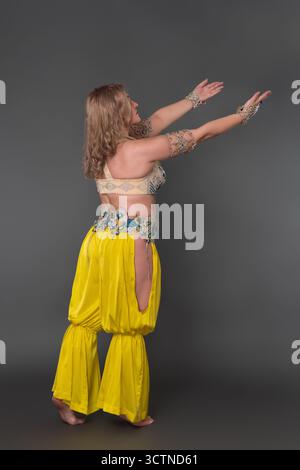 Dreiviertel-Rückansicht einer kurvigen Tänzerin mit Hand erhöht in Tanzkostüm - Oberteil und gelbe Haremshose, bewegt sich barfuß in voller Länge Stockfoto