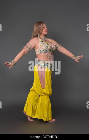 Seitenansicht einer kurvigen Frau mit ausgestreckten Armen, die sich in tanzendem Outfit kleidet - Oberteil und gelbe Haremshose, die barfuß in voller Länge gegen gr Stockfoto