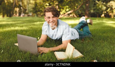 Glücklicher männlicher Student, der einen Laptop benutzt, im Freien online lernt, auf dem Rasen auf dem College-Campus oder im Park liegt Stockfoto