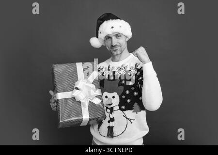 Weihnachtsmann mit Gewinnerschild, Geschenk auf rotem Studiohintergrund, glücklicher junger schwarzer Mann mit Weihnachtsmannmütze und Pullover zu Weihnachten oder Neujahr. Stockfoto