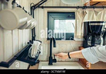 Eine moderne Küche mit elegantem schwarzen Wasserhahn und Kaffeemaschine. Eine Hand greift nach dem Wasserhahn. Weiße Holzwände und hängende Tassen sorgen für eine gemütliche Atmosphäre Stockfoto