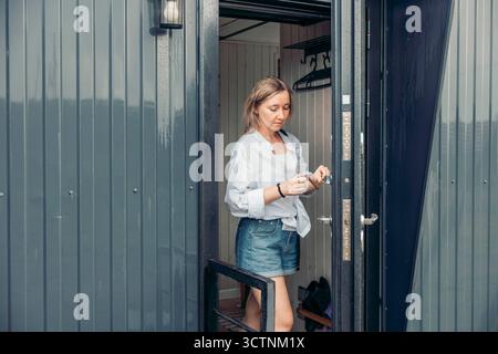 A young Caucasian woman with blonde hair stands at the entrance of a modern cabin. She wears a light blue shirt and denim shorts, holding a smartphone Stockfoto