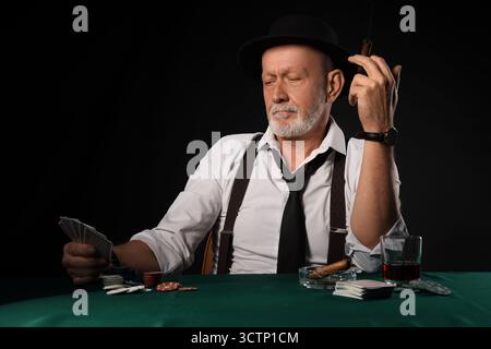 Stilvoller alter Mann mit Zigarre, Spielkarten und Whiskey am Tisch im Casino Stockfoto