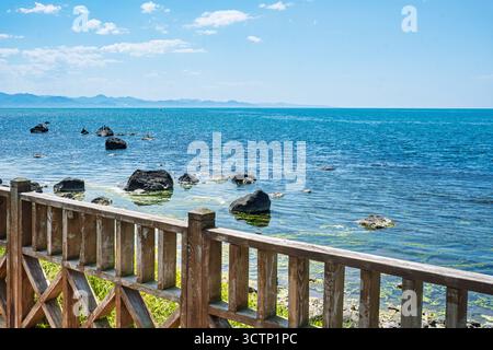 Eine ruhige Landschaft mit dem blauen Wasser eines Meeres. Blick hinter einem rustikalen Holzzaun mit weit entfernten Bergen unter dem Himmel. Stockfoto