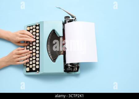Frau tippt auf Schreibmaschine mit leerem Papierbogen auf blauem Hintergrund Stockfoto