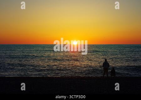 Silhouettenfiguren, die den Sonnenuntergang am Strand beobachten. Eine ruhige Strandszene mit zwei Silhouettenfiguren, die bei Sonnenschein am Wasserrand stehen Stockfoto