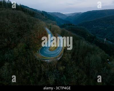 Luftaufnahme einer gewundenen Haarnadelkurve auf einer Bergstraße, die an einem trostlosen, bewölkten Tag durch den dichten Eiffelwald führt. Stockfoto