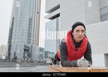 Junge entschlossene Kaukasierin in warmer Sportswear, die Plank macht und Musik hört, während sie im zentralen Geschäftsviertel Outdoor-Workout macht Stockfoto