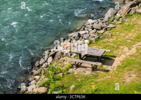 Picknicktisch am Fluss. Ein alter hölzerner Picknicktisch, eingebettet in einen Fluss. Umgeben von glatten Steinen und üppigem Grün. Draufsicht. Stockfoto